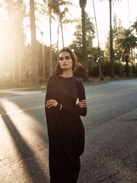 Adult female in long dark dress standing on asphalt road flanked by palm trees with bright backlight and long shadows.