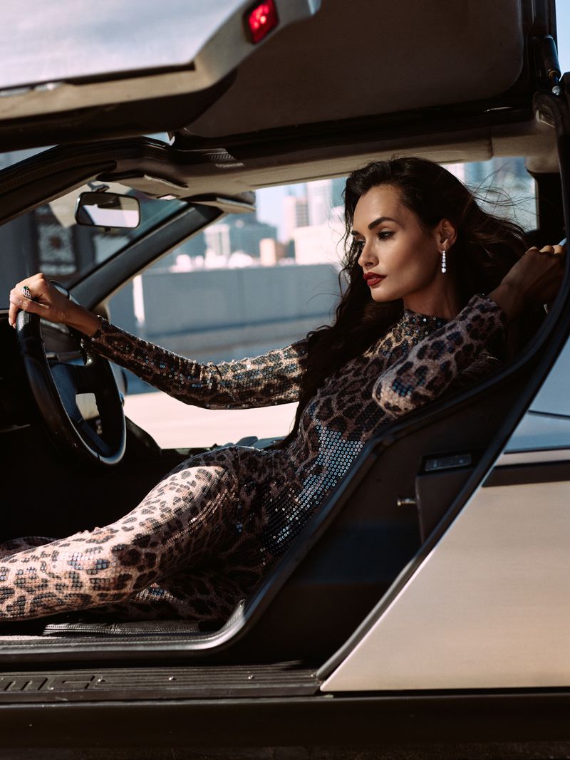 Woman in a cheetah print dress drives a DeLorean car with the door open in an urban scene with tall buildings in the background.