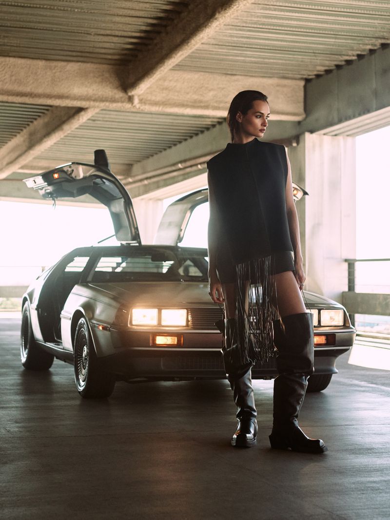 Stylish adult female posing by a classic DeLorean car in a concrete parking garage. Automotive and fashion visual content.