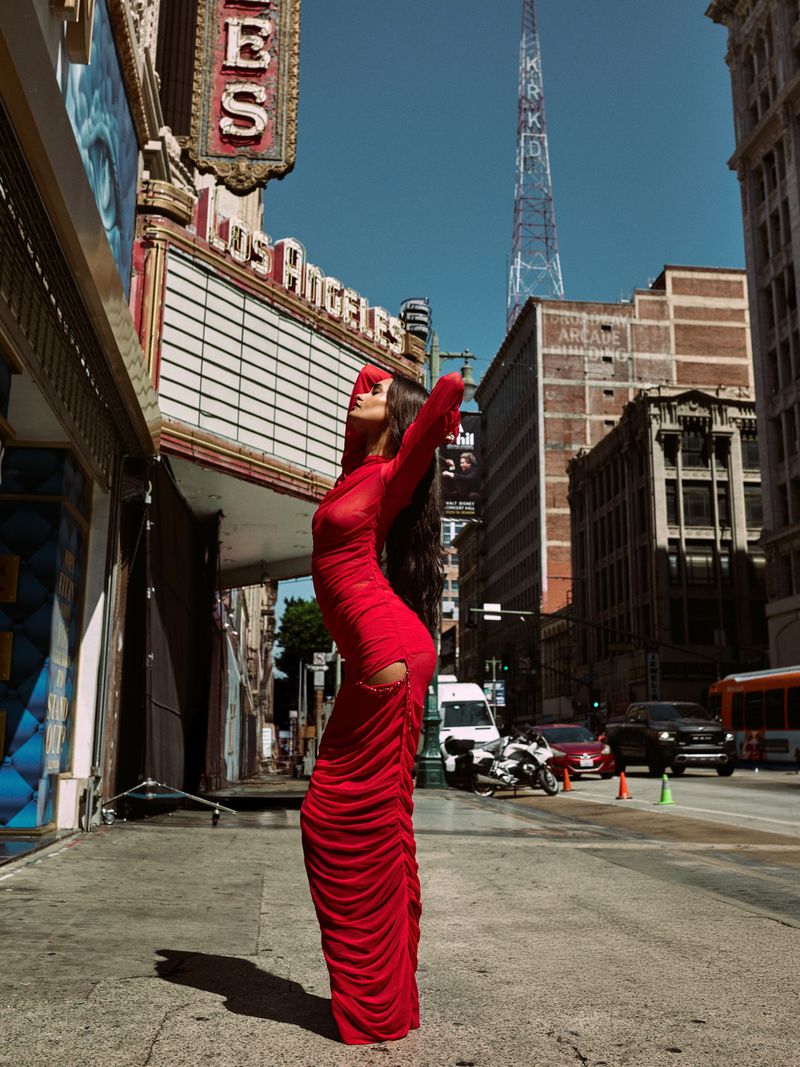 Woman in a red dress poses in front of the Los Angeles Theatre on a city street with buildings and a radio tower in the background.