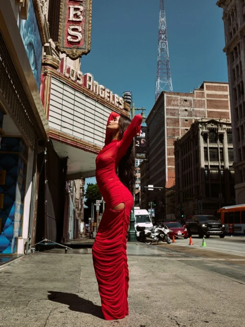 Woman in a red dress poses in front of the Los Angeles Theatre on a city street with buildings and a radio tower in the background.