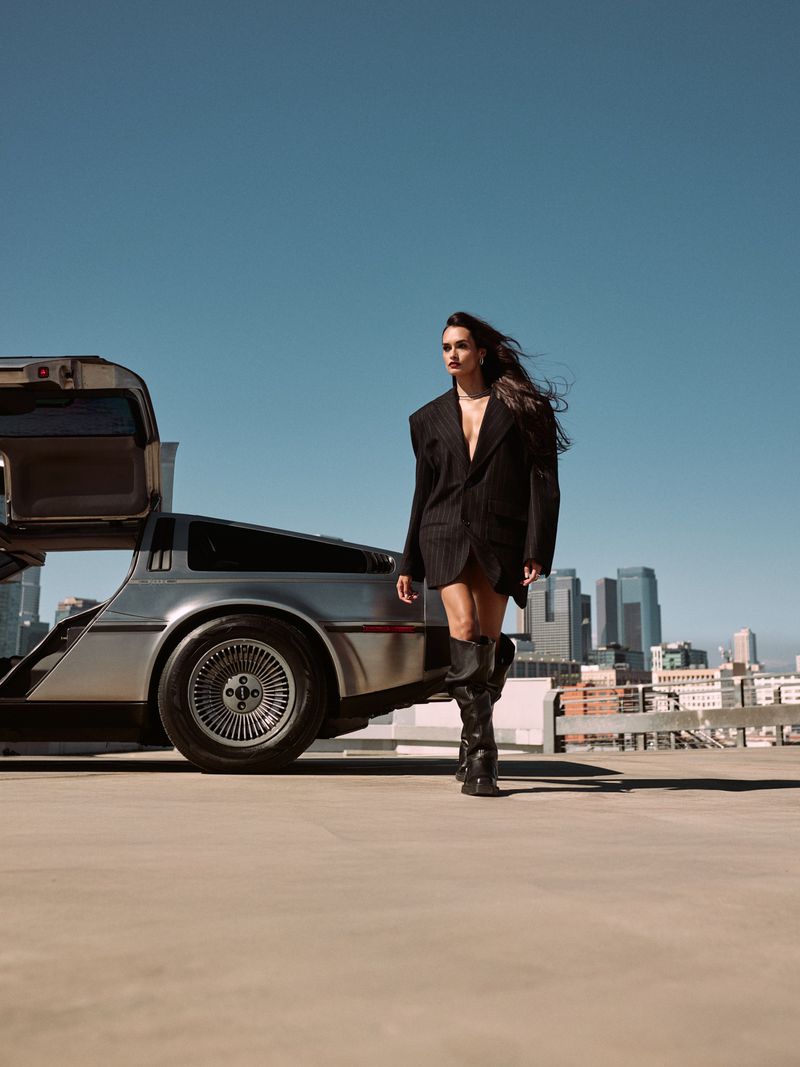 An adult woman poses in an oversized blazer and boots next to a DeLorean on a rooftop with a city skyline backdrop on a clear day.