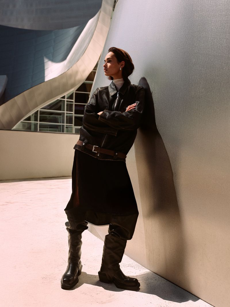 A woman in a black leather jacket and black skirt stands leaning against a concrete wall in an indoor architectural space.