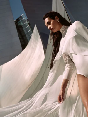 Model Gizele Oliveira in a high-neck white bodysuit and dramatic flowing white cape in front of sculptural architecture.
