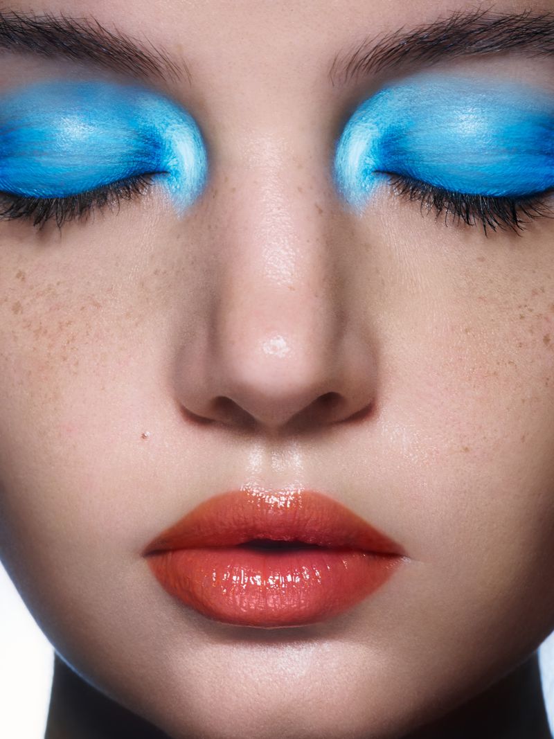 Close-up of a woman's face with vibrant blue eyeshadow and glossy orange lips, emphasizing beauty and cosmetic artistry.