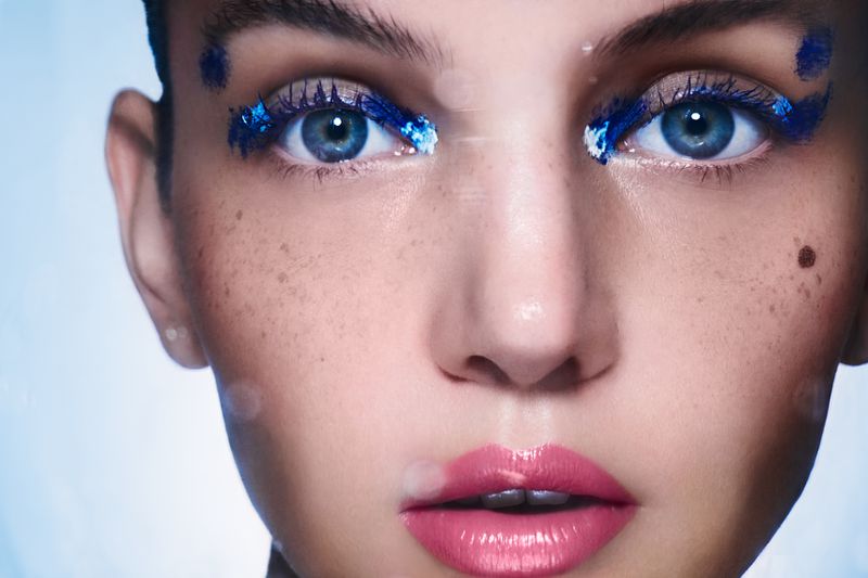 Close-up of a woman's face featuring blue glitter makeup around her eyes and pink glossy lips, set against a light blue backdrop.