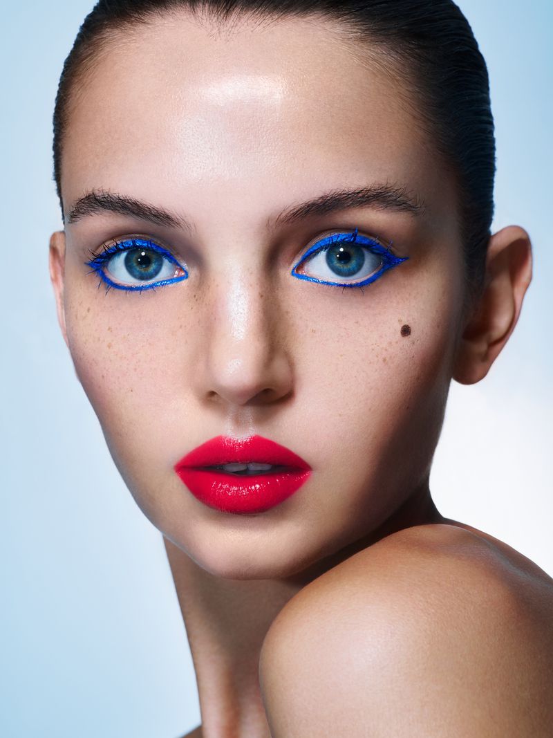 Close-up portrait of a young woman with bright red lipstick and bold blue eyeliner against a pale blue gradient background.