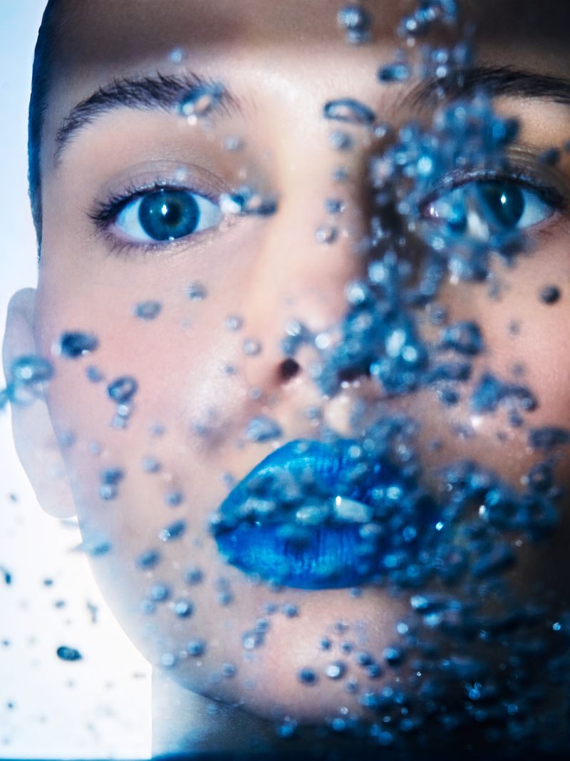 Close-up of a woman's face with blue eyes and blue lips, partially obscured by water droplets against a bright, light blue background.