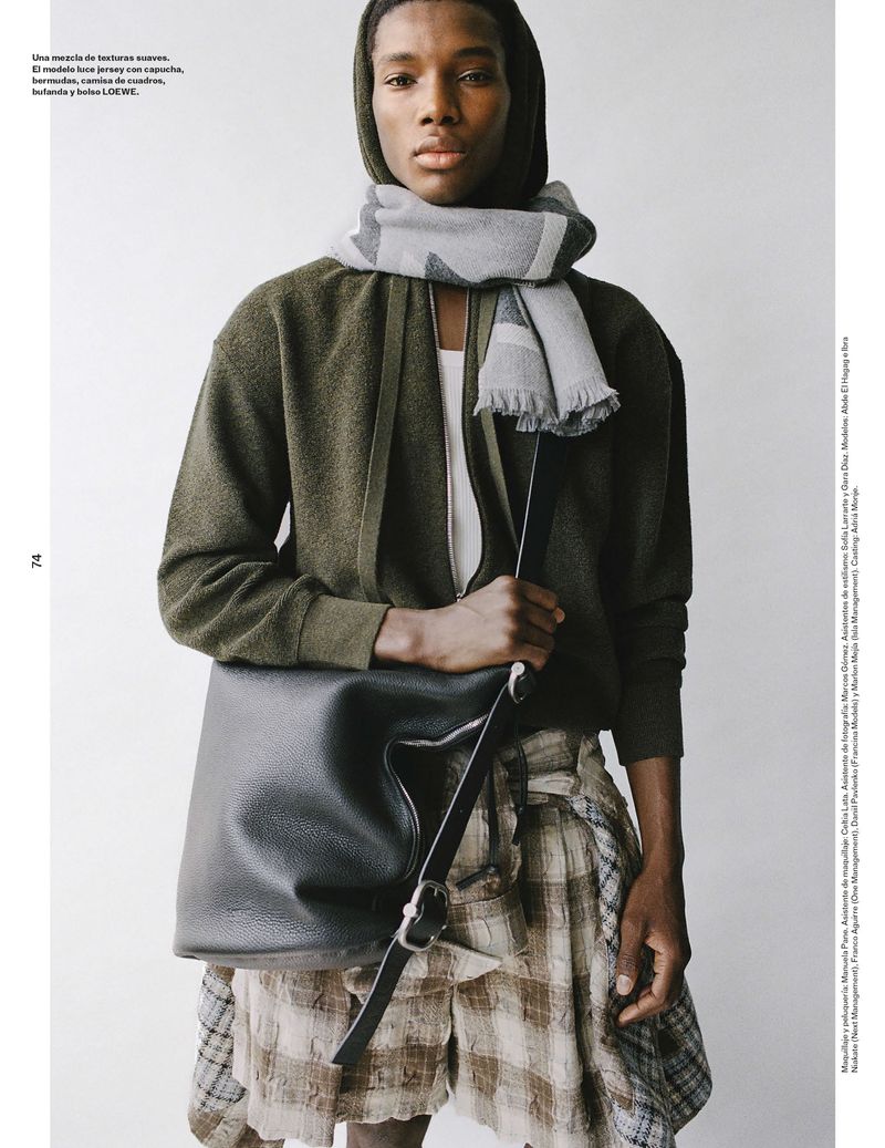 Male fashion model poses in a hooded jacket and plaid shorts with a large leather shoulder bag in a neutral studio.