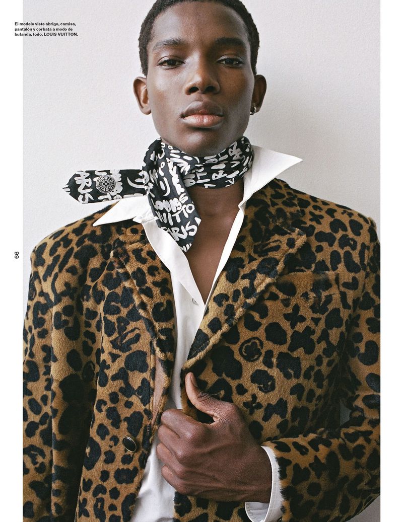 A stylish Black man with a patterned jacket, white shirt, and neck scarf, poses against a neutral backdrop in a fashion editorial shot.