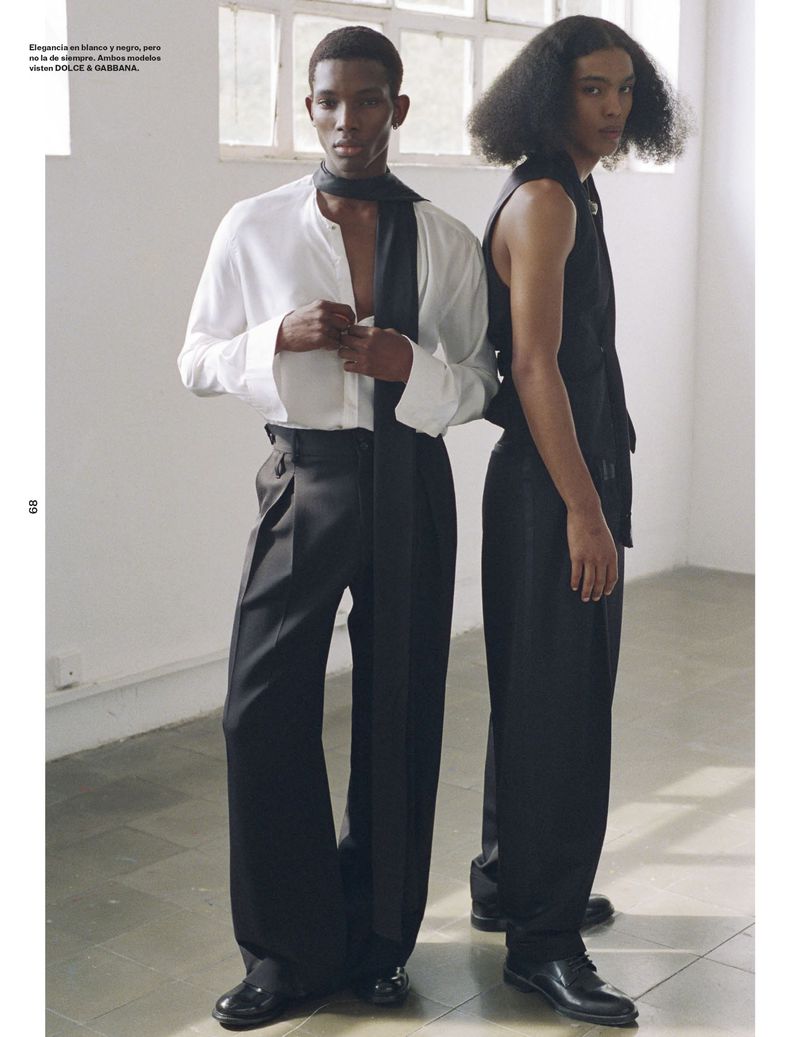 Two dark-skinned male fashion models pose indoors wearing black and white outfits, one in a white shirt and the other in a black vest.