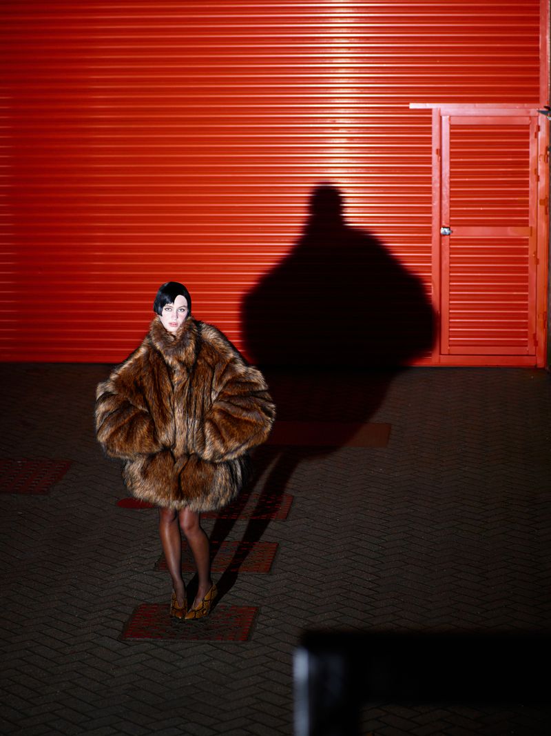 Fashion portrait of a woman in a fur coat standing in front of a red corrugated metal wall with a large shadow. Dark, edgy style.