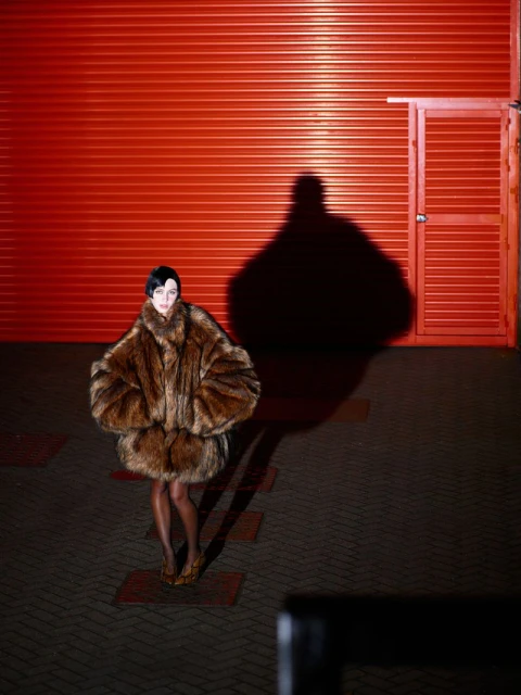Fashion portrait of a woman in a fur coat standing in front of a red corrugated metal wall with a large shadow. Dark, edgy style.