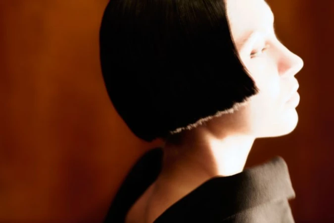 Profile portrait of an adult female with a short black bob haircut, wearing a black garment, against a blurry brown background.