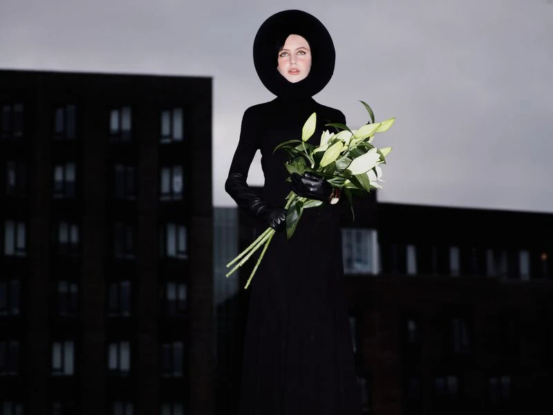 Elegant woman in black ensemble and large hat holds a bouquet of lilies in front of a dark, modern building and overcast sky.