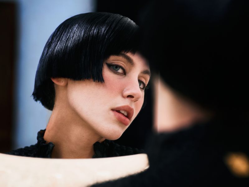Headshot of a woman with a short, blunt black bob hairstyle, light skin, and dark eye makeup, looking into a mirror.