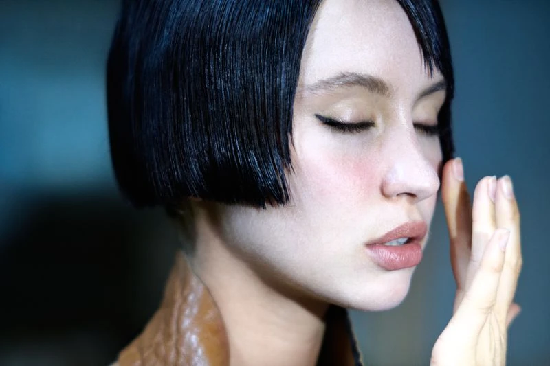 Close-up of a woman with a modern, black bob hairstyle and closed eyes, wearing a brown jacket in a dimly lit indoor setting.