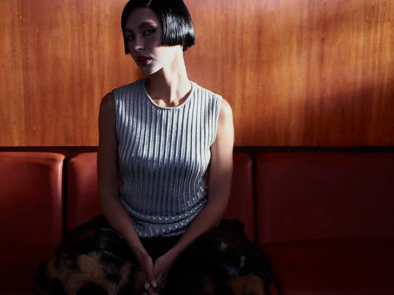 Portrait of a woman with a sleek black bob haircut, wearing a ribbed silver top, seated on a red couch against a wood wall.