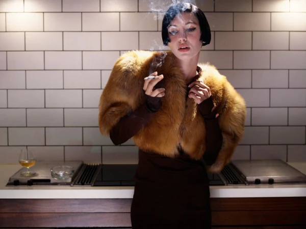 Model Iris Law with dark bob hair and faux fur stole smoking a cigarette in kitchen