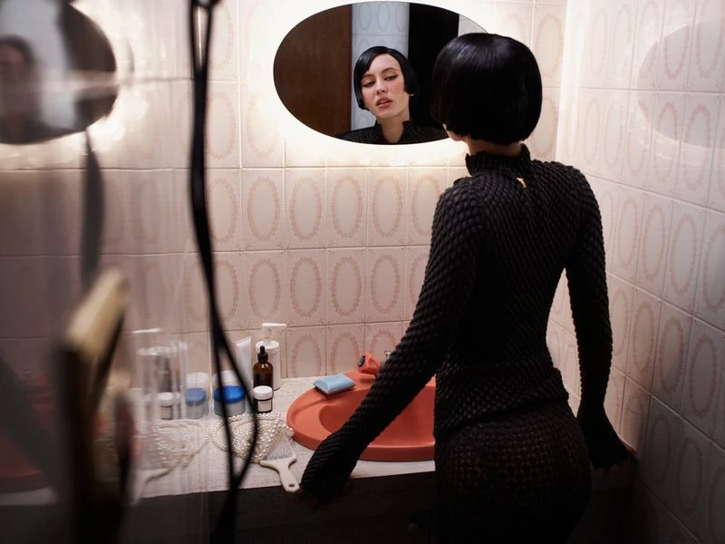 Woman in textured black dress stands in bathroom, gazing into a lit oval mirror with toiletries on sink counter and patterned tile.