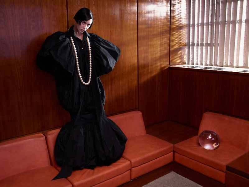 A stylish woman in a black gown poses in an indoor setting with wood-paneled walls, red couches, a glass orb, and dramatic lighting.