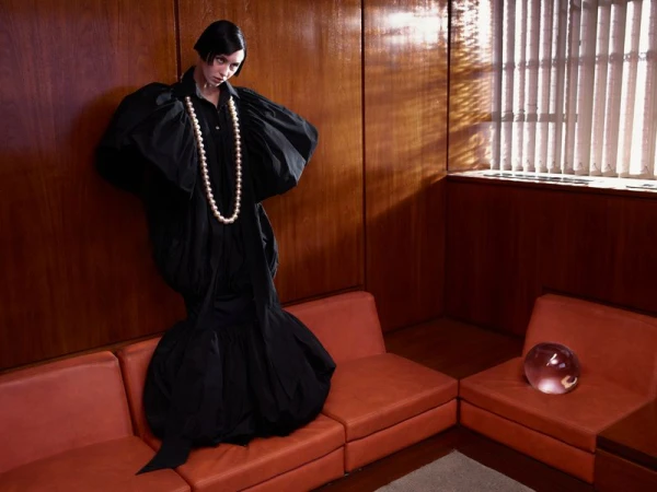 A stylish woman in a black gown poses in an indoor setting with wood-paneled walls, red couches, a glass orb, and dramatic lighting.