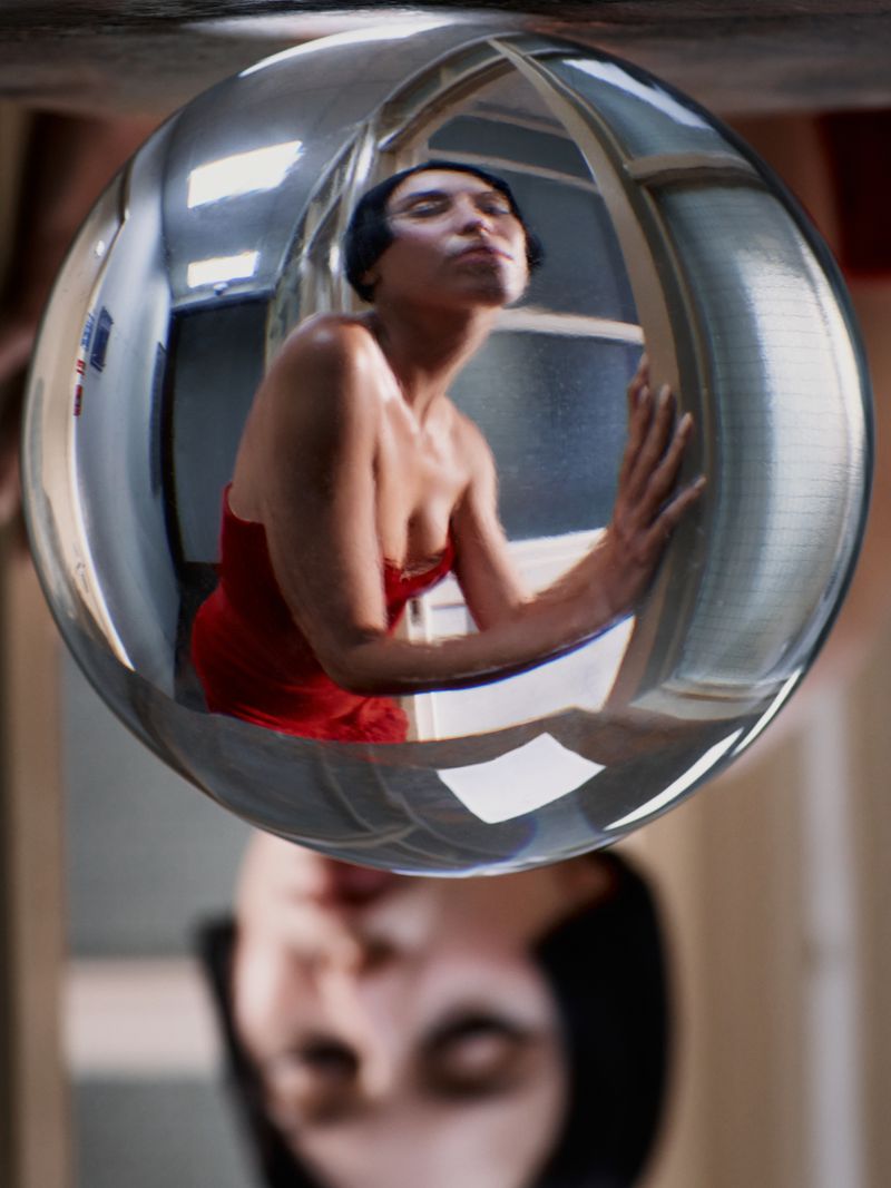 Woman in red dress poses in a reflective sphere, creating a distorted and mirrored image in an indoor setting with window light.