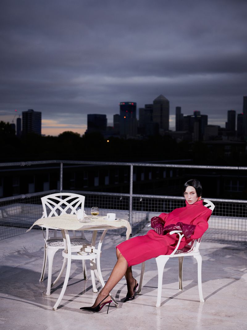 Model Iris Law in a vibrant red dress seated on a rooftop patio at dusk overlooking a distant city skyline
