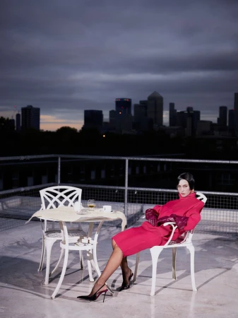Model Iris Law in a vibrant red dress seated on a rooftop patio at dusk overlooking a distant city skyline