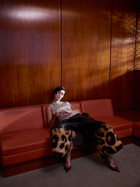 Stylish woman in a sleeveless top and leopard-print pants lounges on an orange couch against a paneled wood wall in a studio setting.