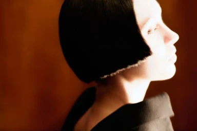 Model Iris Law in profile showing a blunt black bob haircut and dark collar against a warm amber background.
