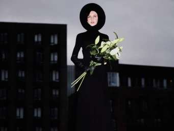Model Iris Law wearing a structured black halo headpiece and black gown holding white lilies before an urban building.