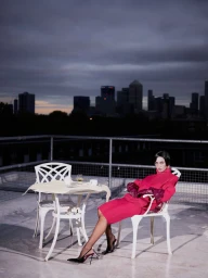 Model Iris Law in a vibrant red dress seated on a rooftop patio at dusk overlooking a distant city skyline