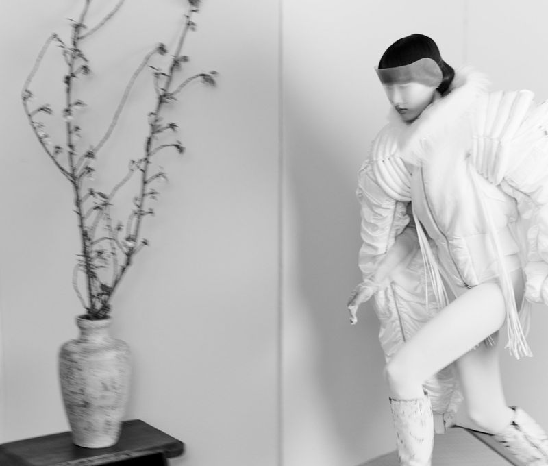 Black and white fashion shot of a woman in avant-garde puffer coat and boots next to a vase with branches on a small wooden table.