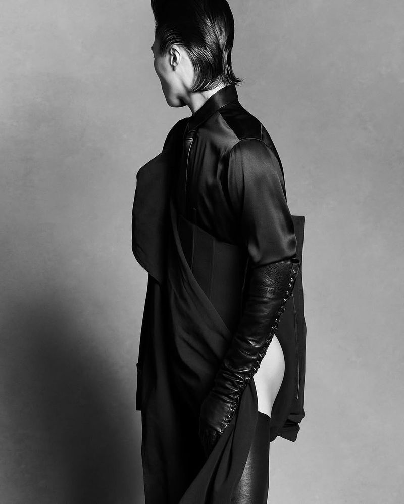 Monochrome studio shot of a person with slicked-back hair wearing a dark, draped garment with long leather gloves, side view against a gray backdrop.