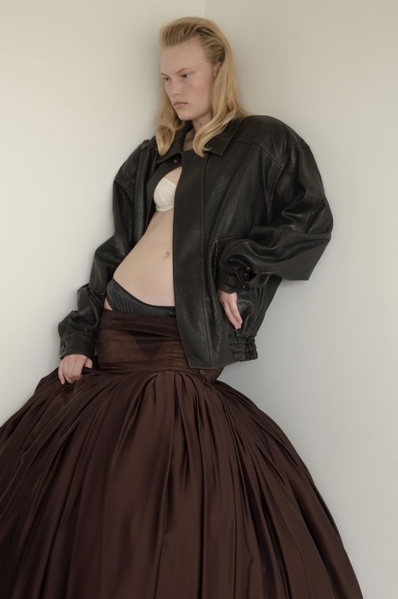 Young woman in black leather jacket and large brown skirt leans against a corner in a fashion photo, showcasing a modern avant-garde style.