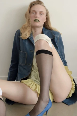 Model seated against a plain wall wearing a blue leather jacket, pale yellow ruffled dress, sheer black knee-highs, and blue heels.