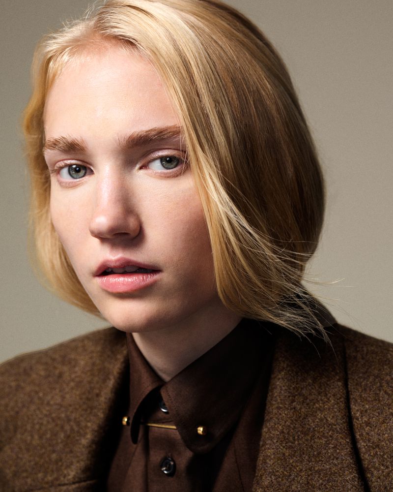 Portrait of a blonde-haired woman in a brown shirt and jacket against a neutral backdrop, focusing on fashion and natural beauty.