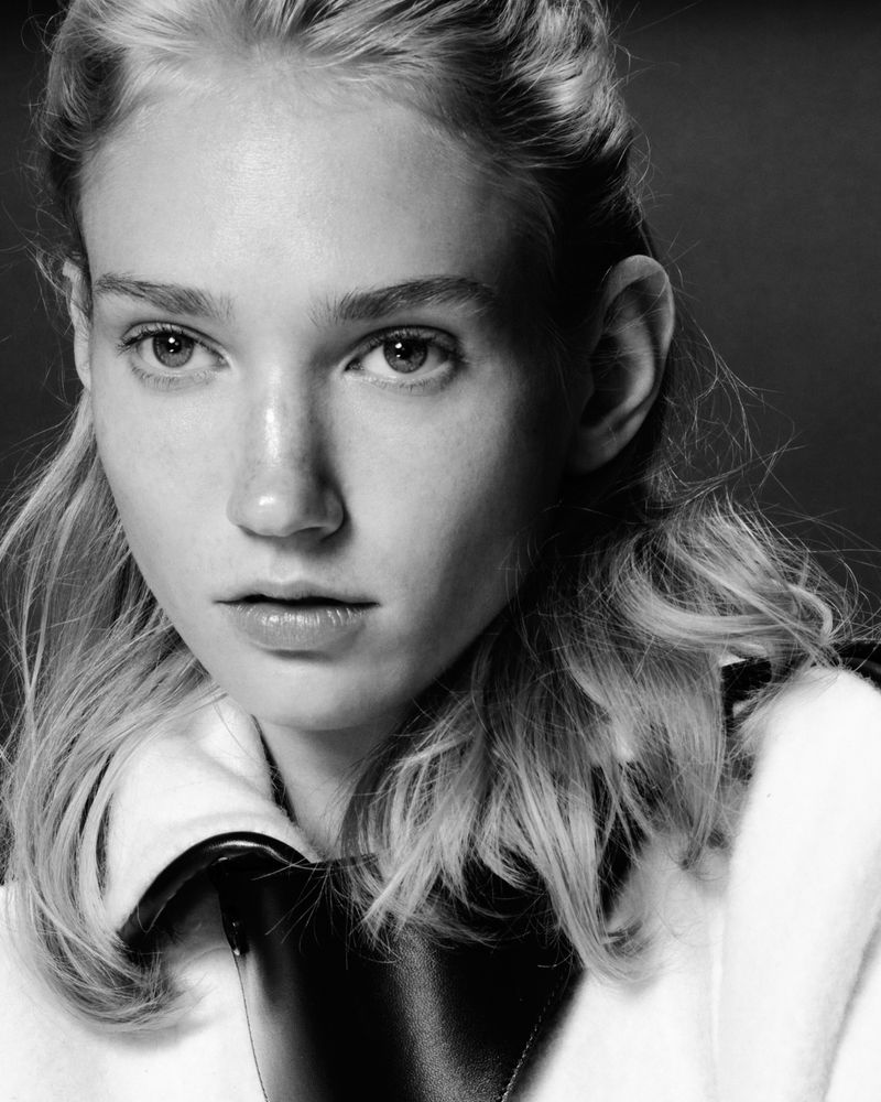Monochrome portrait of a fair-skinned young woman with blonde wavy hair, wearing a white jacket with black trim in a studio.