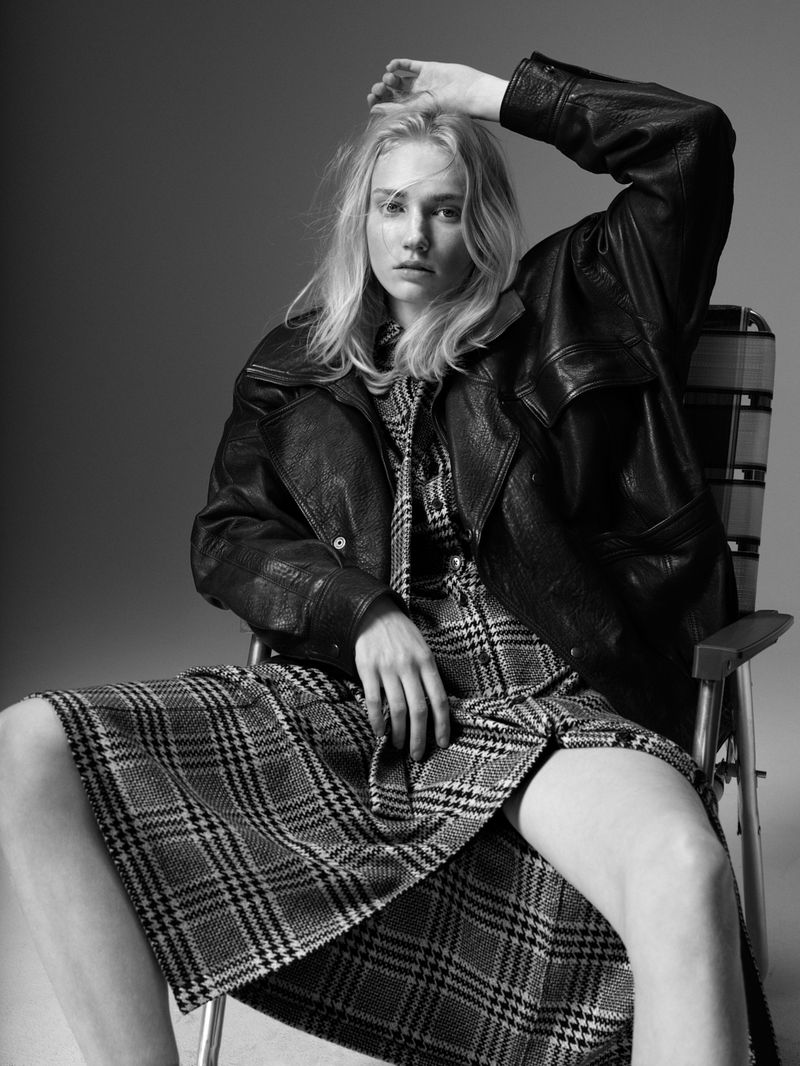 A blonde woman sits in a director's chair in a black and white studio shot, wearing a black leather jacket and a plaid dress.