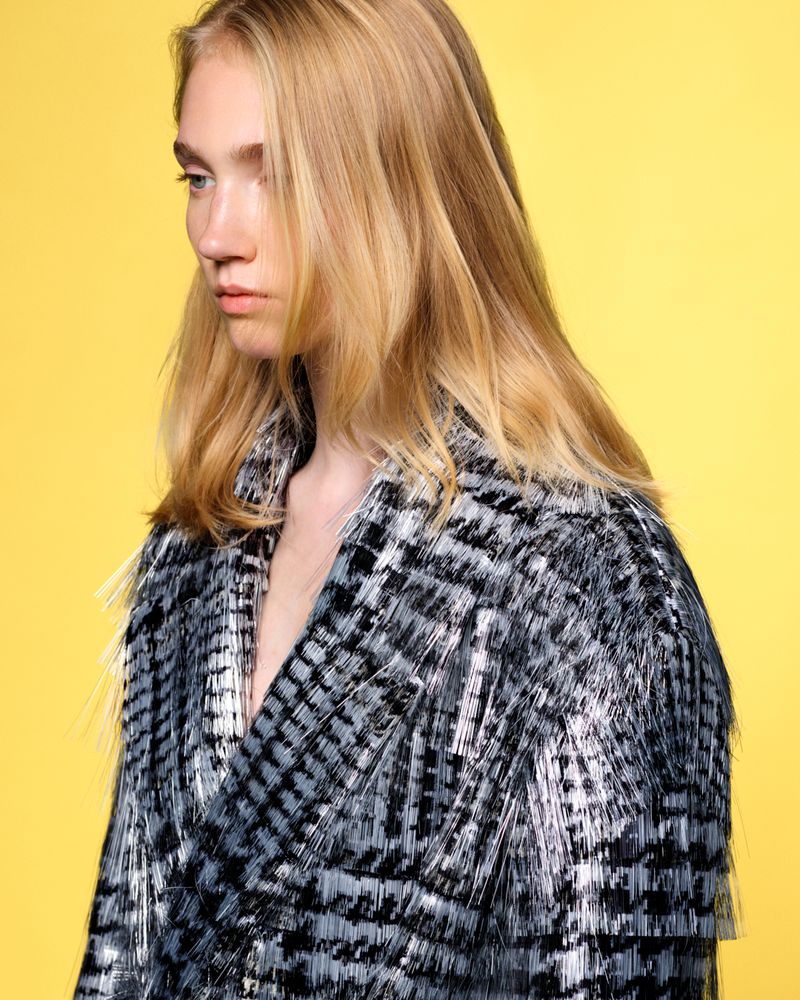Model with blonde hair poses in a studio wearing a black and grey fringed jacket against a solid yellow backdrop for a fashion editorial.