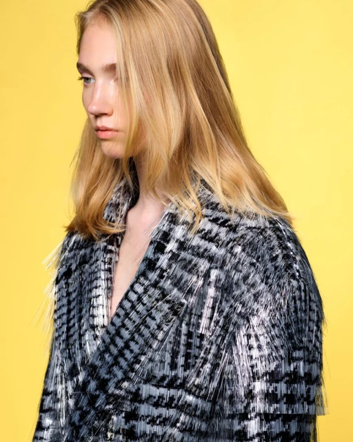 Model with blonde hair poses in a studio wearing a black and grey fringed jacket against a solid yellow backdrop for a fashion editorial.