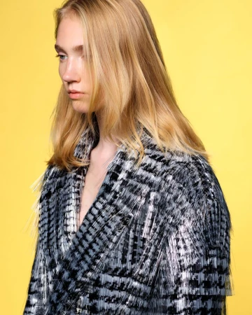 Model with blonde hair poses in a studio wearing a black and grey fringed jacket against a solid yellow backdrop for a fashion editorial.