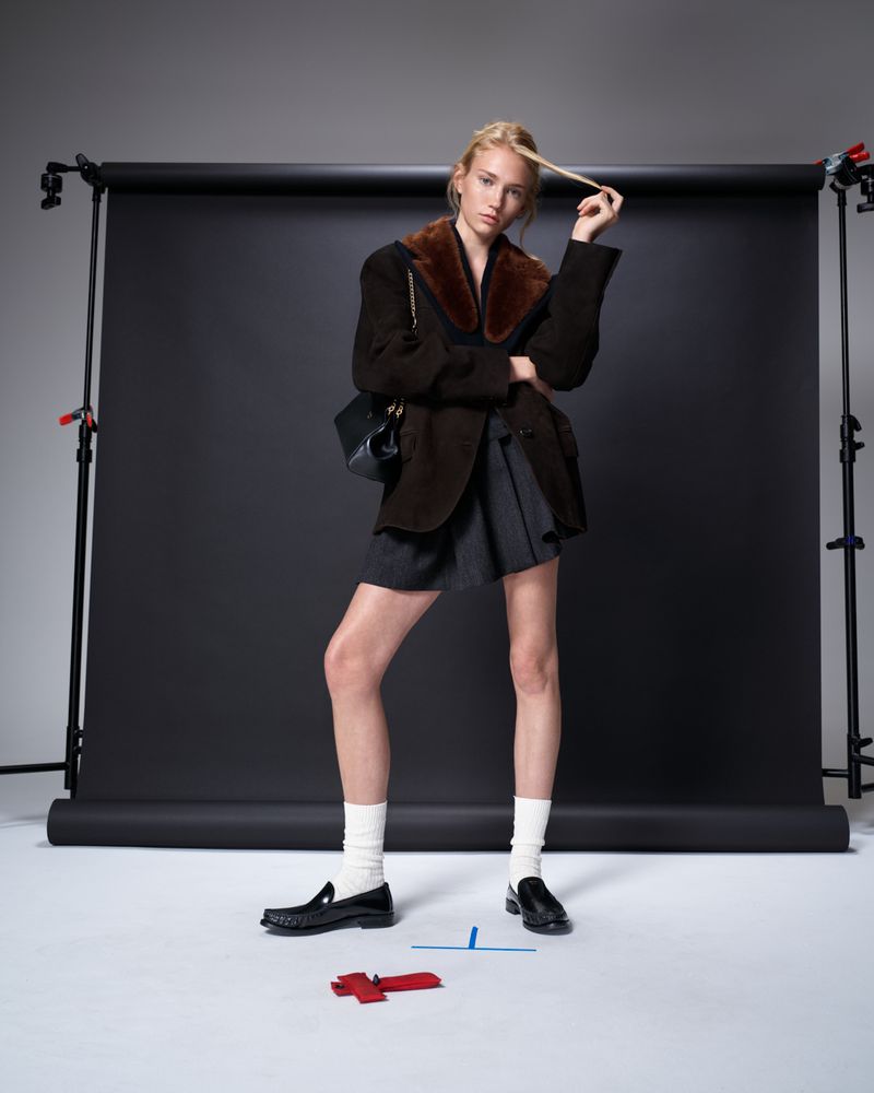 Full-length shot of a woman in a brown blazer, grey skirt, white socks, and black loafers in a photo studio, posing against a black backdrop.