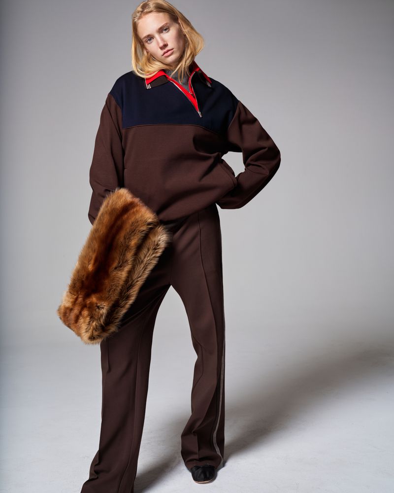 Fashion photo of a blonde model in a brown and navy sweatshirt and pants, holding a fur wrap, posing in a light grey photo studio.