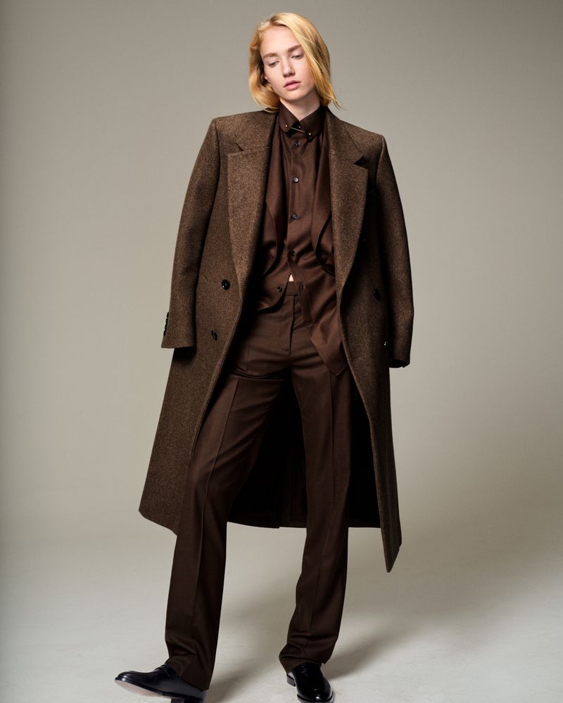 Full-length shot of a person with blond hair wearing a brown suit and long coat in a studio with a light gray background.