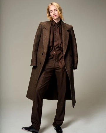 Full-length shot of a person with blond hair wearing a brown suit and long coat in a studio with a light gray background.