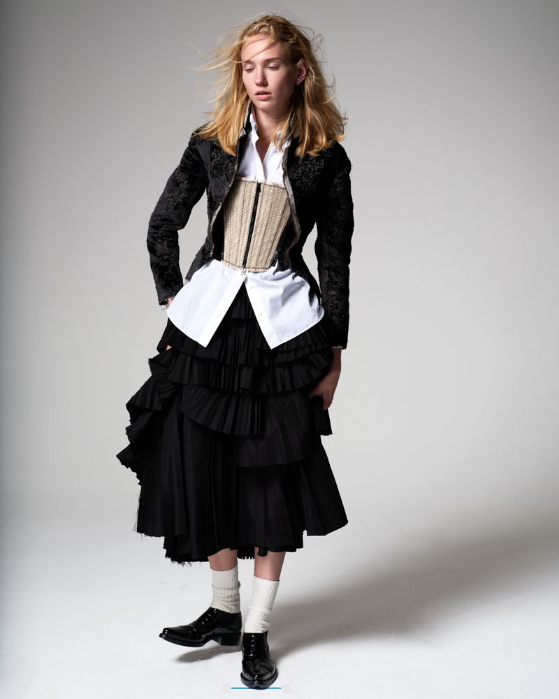 Fashion editorial shot of a female model in a white shirt, beige corset, black jacket, and layered skirt, posing in a photo studio.