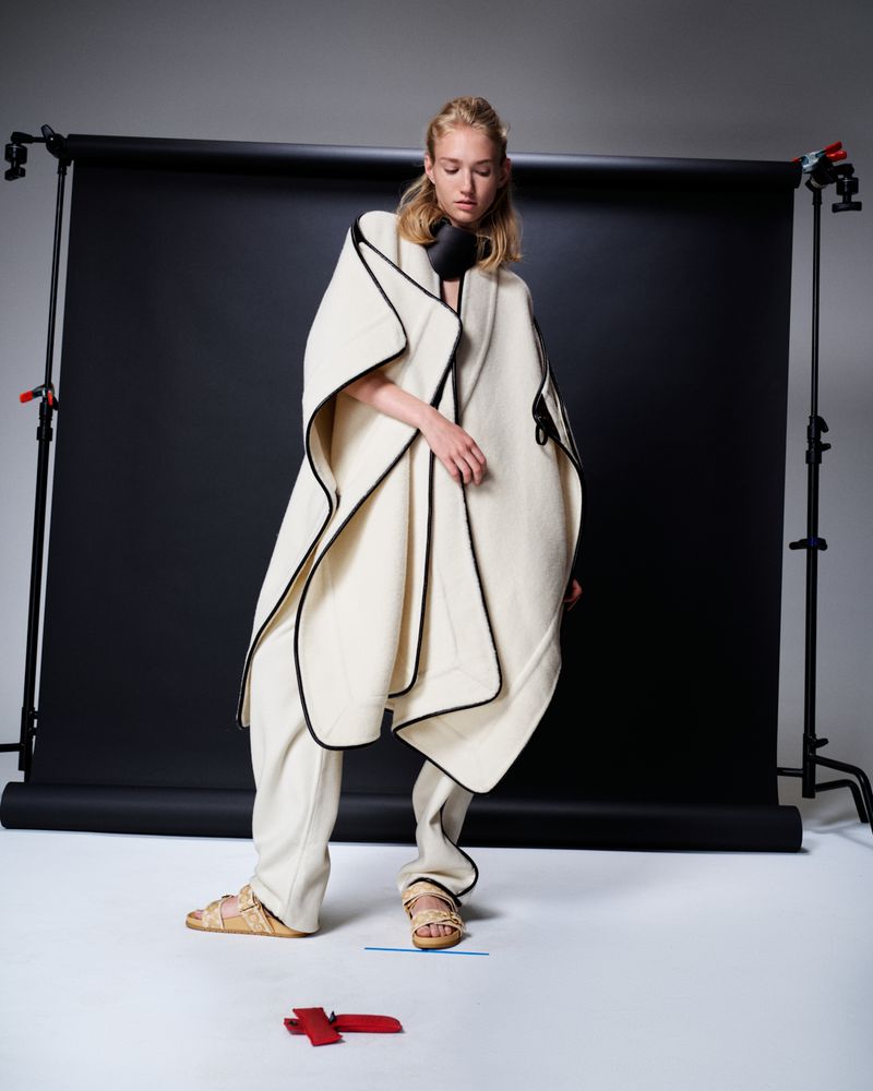 Fashion shot of a blonde woman wearing a cape and loose pants, posed in a photo studio with a gray background and black backdrop.