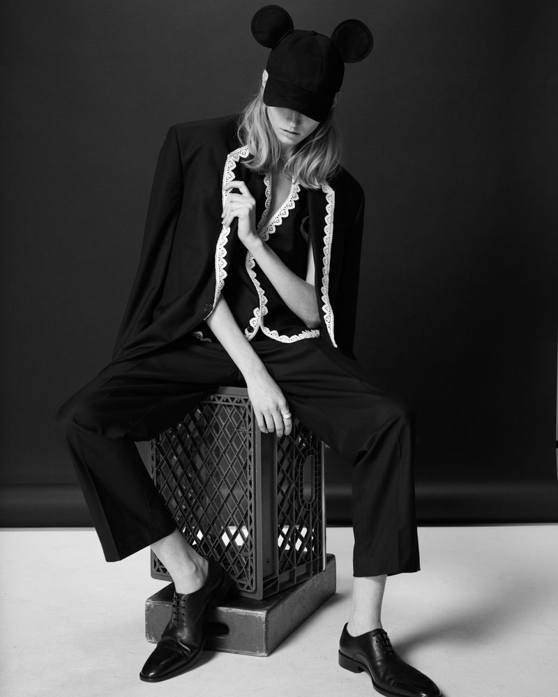 Fashionable woman poses in black suit, blouse with lace trim, and novelty cap, seated on a milk crate against a black background.